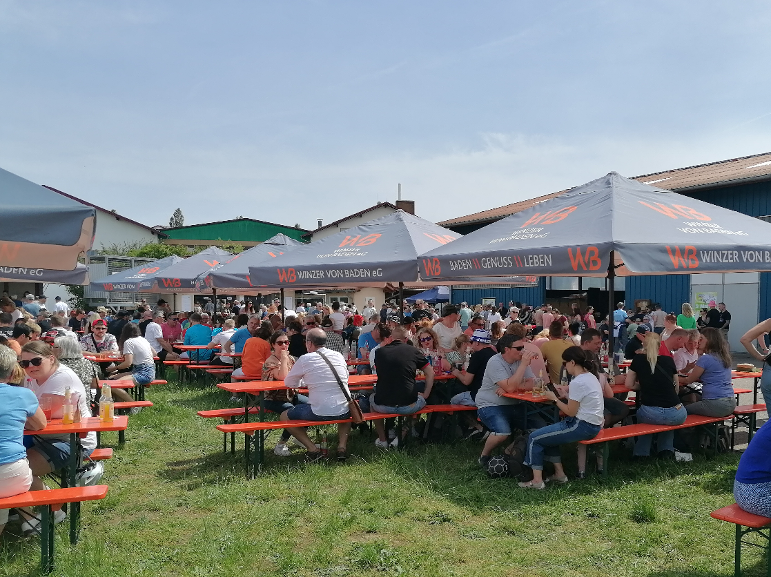 Viele Leute sitzen gesellig unter Sonnenschirmen auf Festzeltgarnituren