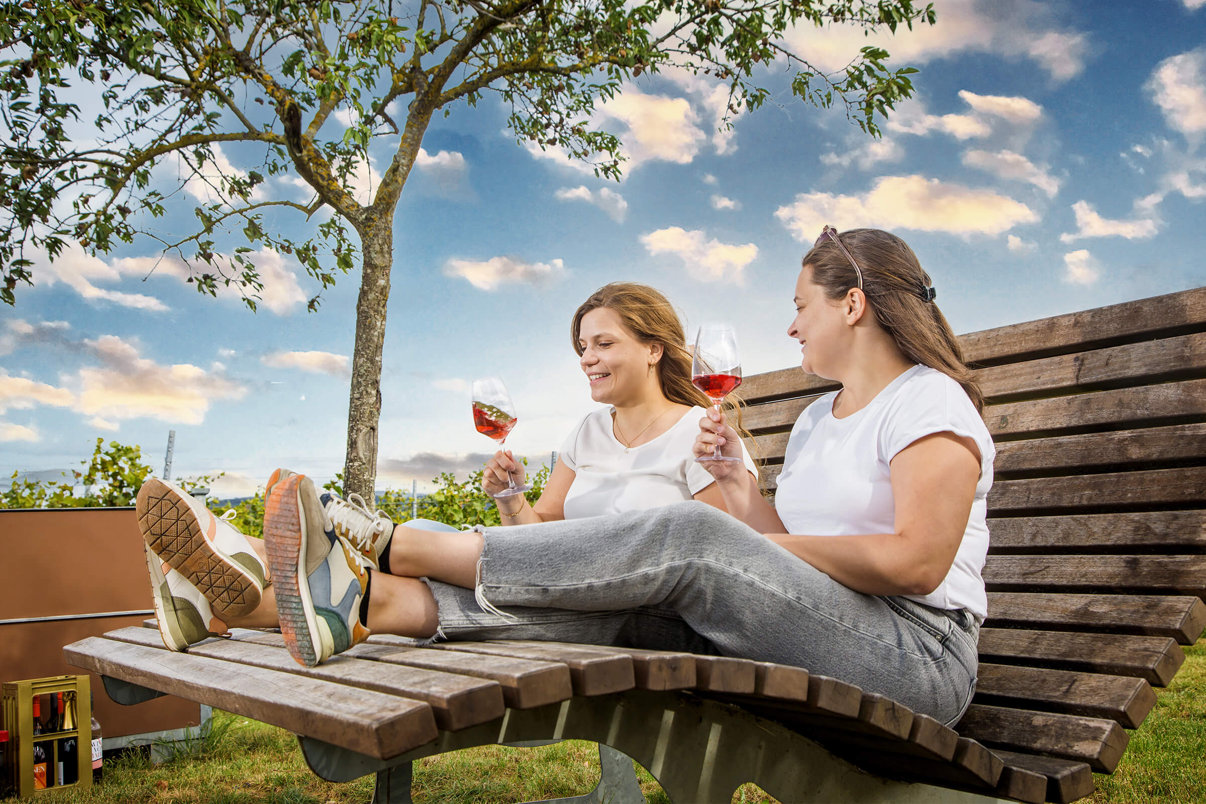 Zwei junge Frauen sitzen auf einer Holzliege im Weinberg und trinken Roséwein